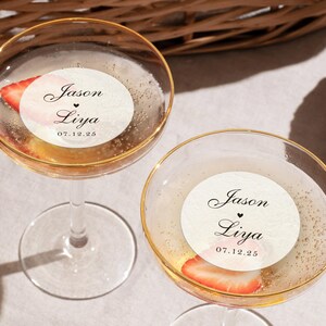 May include: Two champagne glasses with gold rims, filled with a bubbly beverage and a strawberry slice. Each glass has a round sticker with the names "Jason" and "Liya" and the date "07.12.25".