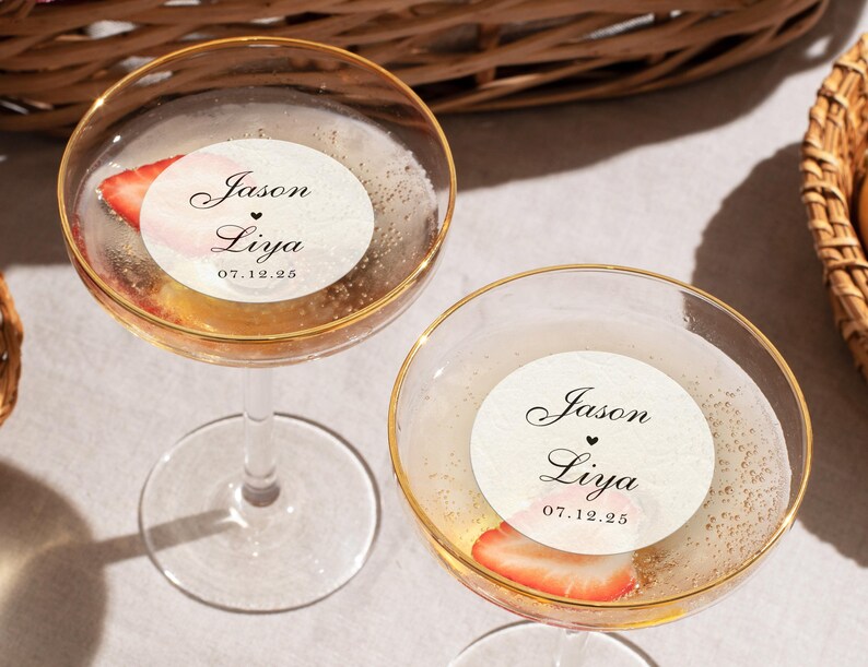 May include: Two champagne glasses with gold rims, filled with a bubbly beverage and a strawberry slice. Each glass has a round sticker with the names "Jason" and "Liya" and the date "07.12.25".