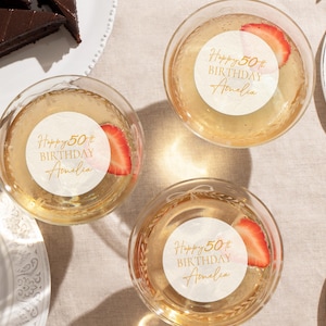 May include: Overhead view of three champagne glasses with strawberry slices. Each glass has a white sticker with gold text reading "Happy 50th Birthday Amelia". Slices of chocolate cake are on white plates.