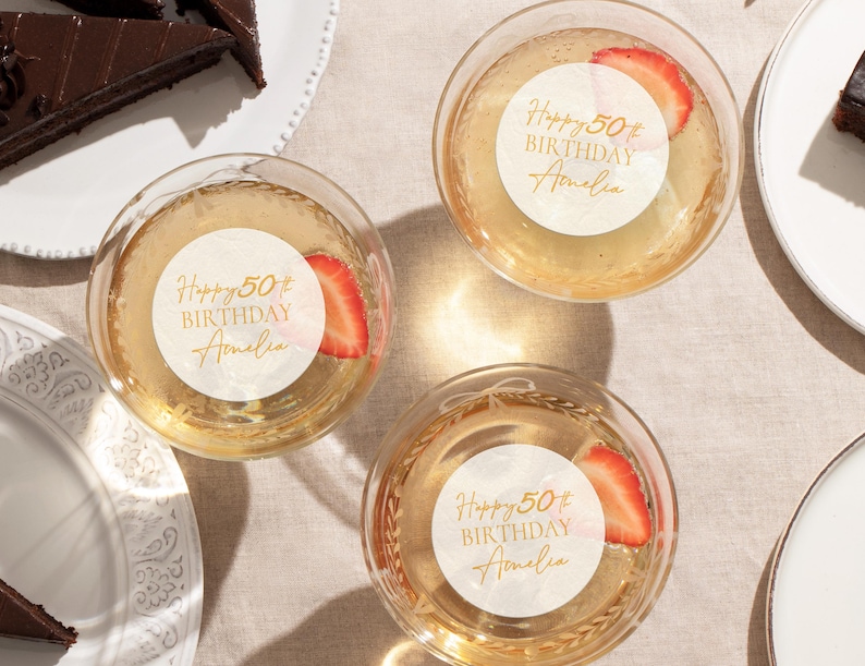 May include: Overhead view of three champagne glasses with strawberry slices. Each glass has a white sticker with gold text reading "Happy 50th Birthday Amelia". Slices of chocolate cake are on white plates.