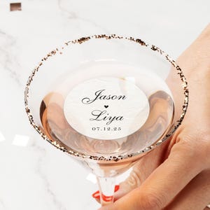 May include: A martini glass with a gold glitter rim, filled with a pink liquid. The glass has a white sticker with the names "Jason" and "Liya" and the date "07.12.25". The glass is held by a person.