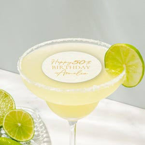 May include: A margarita cocktail in a stemmed glass with a salt-rimmed edge, garnished with a lime wedge. A circular label on the drink reads "Happy 50th Birthday Amelia" in gold script. Sliced limes are on a glass plate.