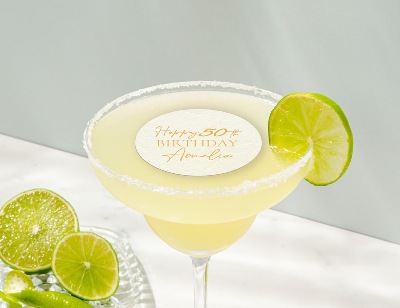 May include: A margarita cocktail in a stemmed glass with a salt-rimmed edge, garnished with a lime wedge. A circular label on the drink reads "Happy 50th Birthday Amelia" in gold script. Sliced limes are on a glass plate.