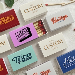May include: A collection of rectangular matchboxes in various colors including red, white, light blue, and pink, featuring printed logos such as Thrashin Supply Co, Heritage Paint Co, Belfast Brunch Co, Tyson's Tacos, and The Hudson Hotel & Cafe.