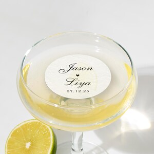 May include: A champagne glass filled with a yellow liquid, possibly a cocktail, with a white circular sticker inside. The sticker has the names "Jason" and "Liya" with a heart symbol and the date "07.12.25". A lime slice is in the foreground.