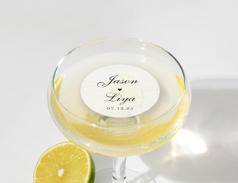 May include: A champagne glass filled with a yellow liquid, possibly a cocktail, with a white circular sticker inside. The sticker has the names "Jason" and "Liya" with a heart symbol and the date "07.12.25". A lime slice is in the foreground.