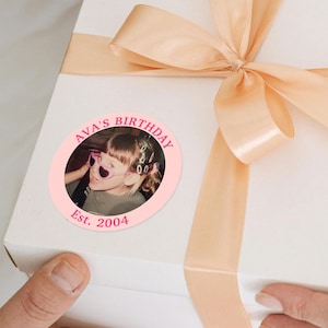 Puede incluir: Una caja de regalo blanca con una cinta y un lazo color melocot&oacute;n. Una pegatina circular rosa en la caja dice "AVA'S BIRTHDAY" alrededor de una foto de un ni&ntilde;o con gafas de sol en forma de coraz&oacute;n. La pegatina tambi&eacute;n dice "Est. 2004".