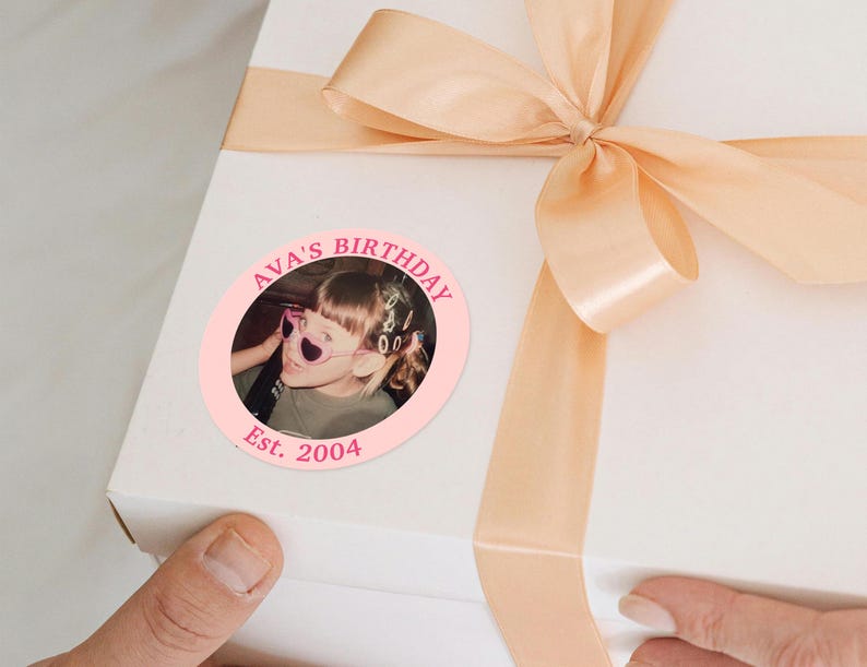 Puede incluir: Una caja de regalo blanca con una cinta y un lazo color melocot&oacute;n. Una pegatina circular rosa en la caja dice "AVA'S BIRTHDAY" alrededor de una foto de un ni&ntilde;o con gafas de sol en forma de coraz&oacute;n. La pegatina tambi&eacute;n dice "Est. 2004".