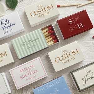 May include: A collection of rectangular matchboxes featuring various custom designs, including names, dates, and monograms in gold, blue, red, and green ink. One open matchbox reveals wooden matches with red tips. The boxes are arranged on a textured surface.