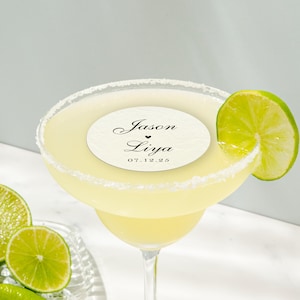 May include: A margarita cocktail in a stemmed glass with a salt-rimmed edge, garnished with a lime wedge. A circular label with the names "Jason & Liya" and the date "07.12.25" is on top of the drink. Sliced limes are on the side.