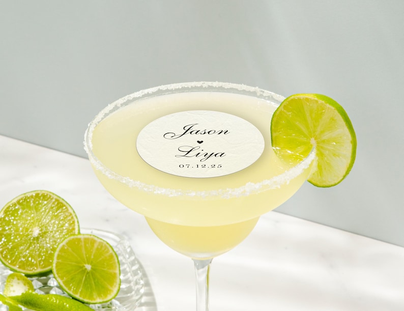 May include: A margarita cocktail in a stemmed glass with a salt-rimmed edge, garnished with a lime wedge. A circular label with the names "Jason & Liya" and the date "07.12.25" is on top of the drink. Sliced limes are on the side.