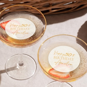 May include: Two champagne glasses with gold rims, filled with a bubbly beverage and a strawberry slice. Each glass has a white sticker with gold text that reads "Happy 50th Birthday Amelia."