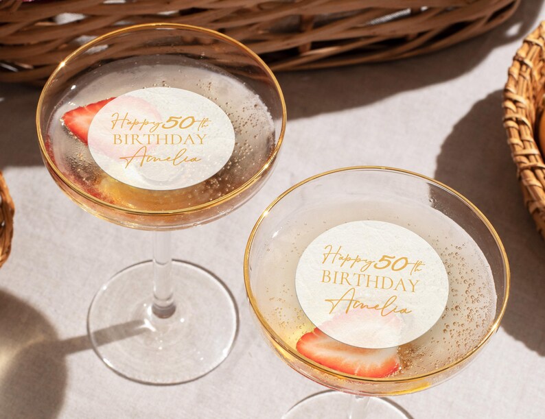 May include: Two champagne glasses with gold rims, filled with a bubbly beverage and a strawberry slice. Each glass has a white sticker with gold text that reads "Happy 50th Birthday Amelia."