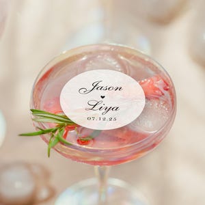 May include: A cocktail in a stemmed glass with ice, fruit, and a sprig of rosemary. A white oval sticker on the glass reads "Jason &hearts; Liya" and "07.12.25" in black script. The drink has a pink hue.