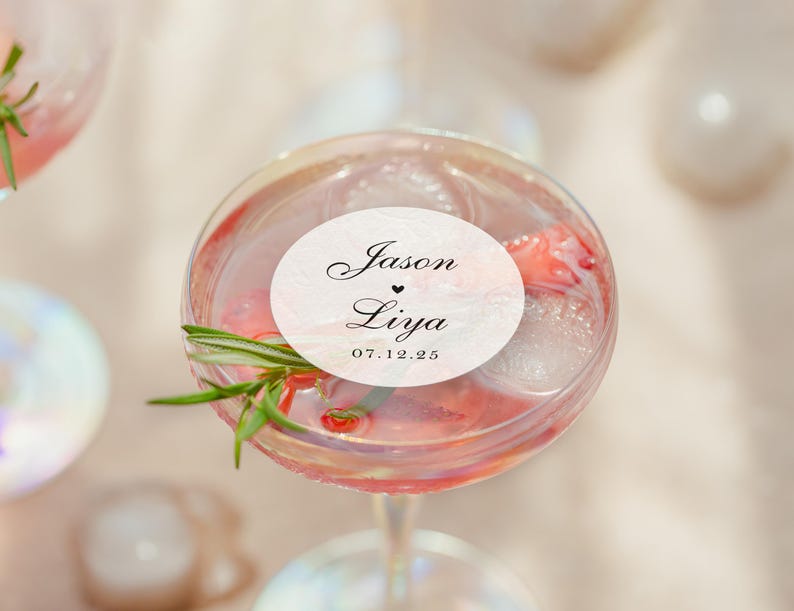 May include: A cocktail in a stemmed glass with ice, fruit, and a sprig of rosemary. A white oval sticker on the glass reads "Jason &hearts; Liya" and "07.12.25" in black script. The drink has a pink hue.
