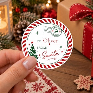 May include: A round white gift tag with a red and white twisted rope border. The tag features a green Christmas tree, red stars, and a North Pole Express postmark stamp. The text reads TO: Oliver and FROM: Santa in red and green lettering.