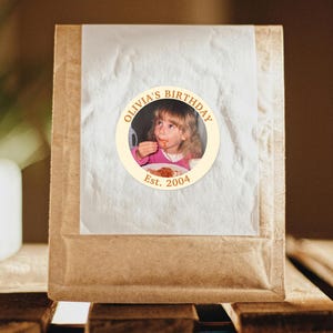 Puede incluir: Un paquete de papel marr&oacute;n con una pegatina circular que muestra una foto de una ni&ntilde;a comiendo. La pegatina dorada dice "Olivia's Birthday" y "Est. 2004". El paquete est&aacute; sellado con papel blanco transl&uacute;cido.