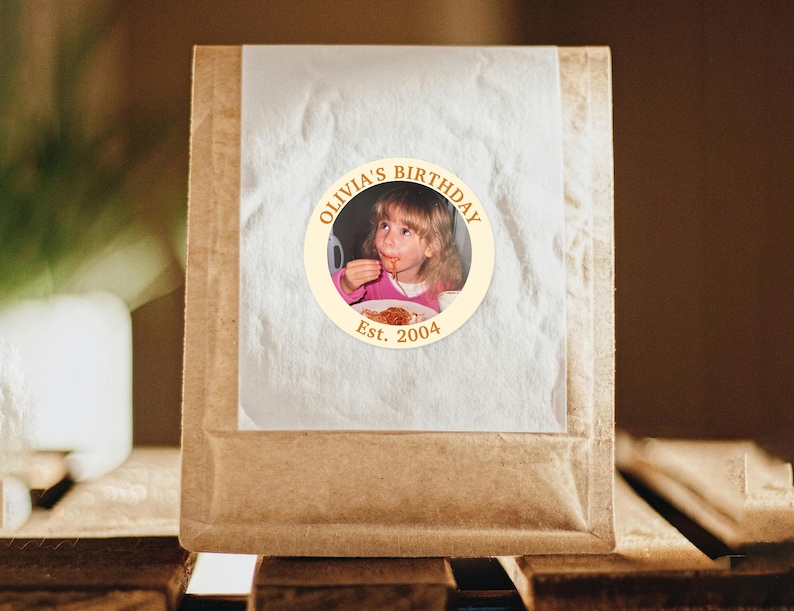 Puede incluir: Un paquete de papel marr&oacute;n con una pegatina circular que muestra una foto de una ni&ntilde;a comiendo. La pegatina dorada dice "Olivia's Birthday" y "Est. 2004". El paquete est&aacute; sellado con papel blanco transl&uacute;cido.