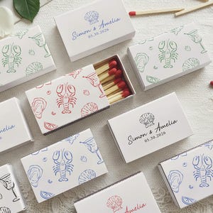 May include: A collection of rectangular matchboxes featuring white covers with printed patterns of lobsters, seashells, and cocktails in red, blue, green, or black ink. Some boxes display the text Simon & Amelia 05.30.2026. One box is open, revealing wooden matches with red tips.
