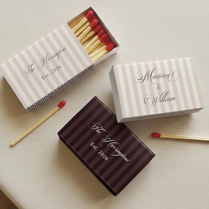 May include: Three rectangular matchboxes with red-tipped wooden matches. Two boxes feature light gray and white vertical stripes with text reading The Harringtons Est. 2026 and Madison & William. One dark brown box has vertical stripes and the same text.