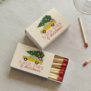 May include: Two rectangular matchboxes with a cream background featuring a yellow taxi carrying a decorated Christmas tree on its roof. The text A NEW YORK Christmas is printed in red. One box is closed, and the other is open, revealing wooden matches.