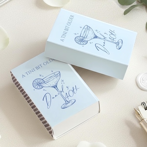 May include: Two light blue matchboxes with a cocktail illustration and the text "A tini bit older" and "Daniel's 40th". The matchboxes are on a white surface.
