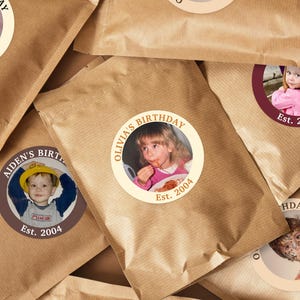 Puede incluir: Bolsas de papel marr&oacute;n con pegatinas circulares con fotos de ni&ntilde;os y el texto "Cumplea&ntilde;os" y "Est. 2004". Las pegatinas tienen un borde marr&oacute;n o granate.