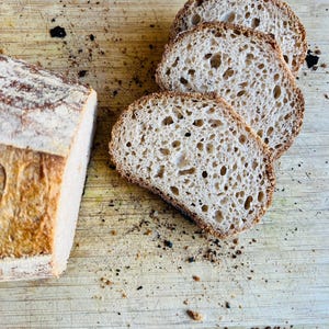 May include: A loaf of freshly baked bread, partially sliced, with three slices arranged on a wooden cutting board. The bread has a golden-brown crust and a porous interior, with visible air pockets. The cutting board has dark spots.