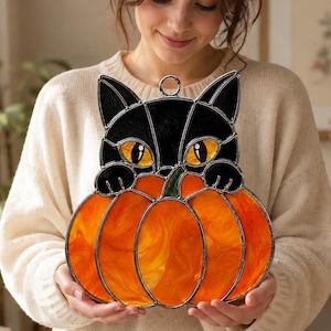 Op de afbeelding: Een glas-in-lood suncatcher in de vorm van een zwarte kat die over een grote oranje pompoen kijkt. De kat heeft heldergele ogen en zwarte oren, terwijl de pompoen is opgebouwd uit gestructureerde oranje glazen segmenten met een groen steeltje.