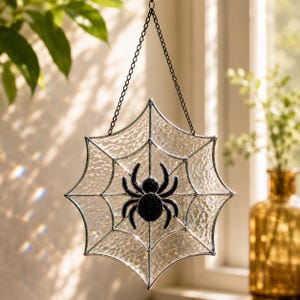 May include: A stained glass suncatcher featuring a clear, textured spider web design with a solid black spider at the centre. The piece is framed in thin metal and hangs from a black metal chain, creating a decorative hanging ornament.