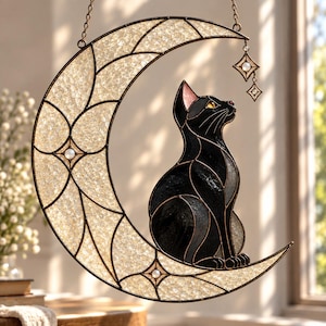Puede incluir: Un atrapasoles de vidrieras que muestra un gato negro sentado sobre una luna creciente. La luna está hecha de vidrio crema texturizado y translúcido con bordes de metal oscuro y pequeñas cuentas blancas. Una estrella cuelga de la luna.