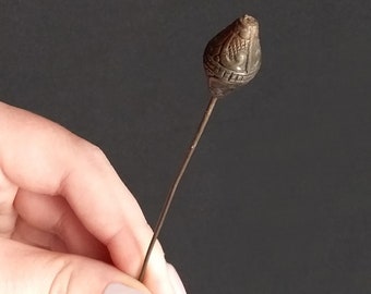 May include: A long, thin, metal pin with a decorative, ornate, bronze-colored head. The head is shaped like a teardrop and has intricate designs.