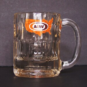 May include: A clear glass mug with a handle. The mug has a raised design on the bottom and a raised design on the top. The mug has a brown and orange A&W logo with a map of the United States in the background.