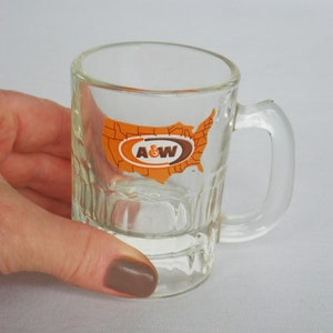 May include: Clear glass shot glass with a handle, featuring the A&W logo inside an orange map of the United States. The glass has a textured design and is held by a hand.