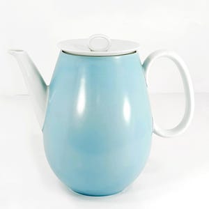 May include: A light blue ceramic teapot with a smooth, rounded body, a white spout, a white handle, and a white lid featuring a small, circular top handle. The teapot has a clean, minimalist design with a glossy finish and a solid color scheme.