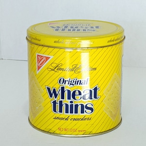 May include: A yellow metal tin with a red and blue Nabisco logo. The tin contains a limited edition of Original Wheat Thins snack crackers. The text on the tin reads "Limited Edition Original Wheat Thins snack crackers, Net Wt 13 oz (369 grams)".