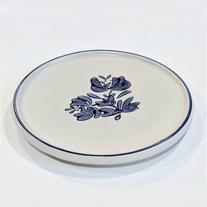 May include: A white ceramic plate with a blue floral design. The plate has a thin blue rim.