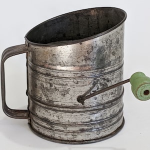 May include: Vintage metal flour sifter with a green wooden handle. The sifter has a handle and a crank to operate it.