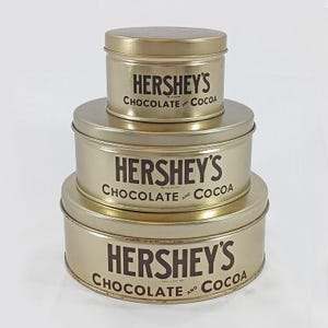 May include: Three gold-colored round metal tins with the text "Hershey's Chocolate and Cocoa" printed in black on each tin.