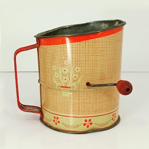 May include: Vintage metal flour sifter with a red handle and trim. The sifter has a cream-colored background with a grid pattern and floral accents. A wooden knob is attached to the side for sifting. The sifter measures approximately 6 inches tall.