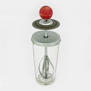 May include: A vintage glass and metal hand-crank milkshake maker with a red knob on top. The glass jar has a metal lid and a metal whisk inside.