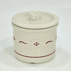 May include: Cream-colored ceramic jar with a matching lid. The jar features a decorative design of maroon floral accents and curved lines. The jar is cylindrical and has a round lid with a small handle. The design is simple and classic.