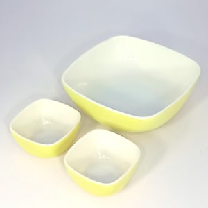 May include: Three nesting bowls with a square shape and rounded corners. The bowls have a bright yellow exterior and a white interior. The largest bowl is in the background, with two smaller bowls in front.