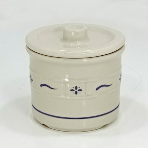 May include: A cream-colored ceramic jar with a matching lid. The jar features a blue floral design and a blue band around the base. The lid has a small handle. The jar is likely used for storage.