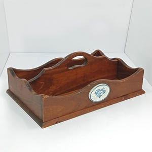 May include: A wooden serving tray with a dark brown finish. The tray has a scalloped edge and a handle. An oval ceramic tile with a blue floral design is affixed to the front. The tray is designed with multiple compartments.