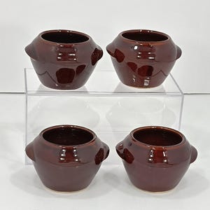 May include: Four small, brown ceramic bowls with rounded bodies and two side handles. The bowls have a glossy finish and are arranged on a clear, tiered surface. The interior of each bowl is dark, contrasting with the exterior.