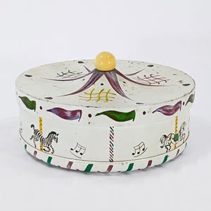 May include: White metal round tin with a carousel design. The tin has a yellow knob on the lid and features a colorful carousel with horses, musical notes, and flags.