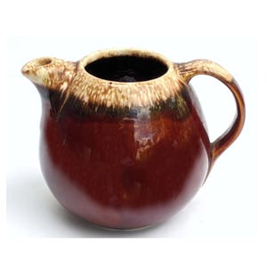 May include: A brown and cream glazed ceramic creamer with a rounded body and a handle. The creamer has a brown glaze with a cream colored rim and a brown glaze on the handle.