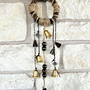 May include: A rustic, circular wall hanging featuring a woven twig base wrapped with twine. Decorative elements include small brass and bronze bells, dark wooden beads, and ornate metal charms, suspended from the base.