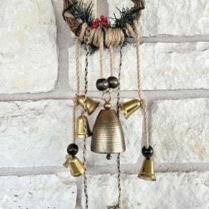 May include: A rustic wall hanging featuring a woven wreath with faux greenery and red berries. Below, a series of brass bells of varying sizes hang from jute twine, creating a charming, decorative piece. The largest bell is approximately 4 inches tall.